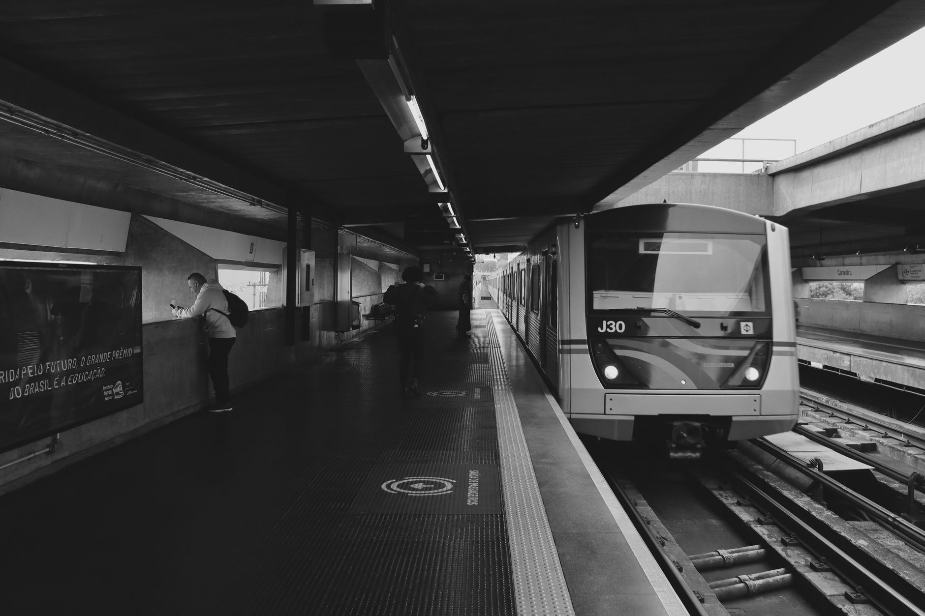 Imagem em preto e branco de uma estação de metrô com um trem chegando e passageiros à vista.