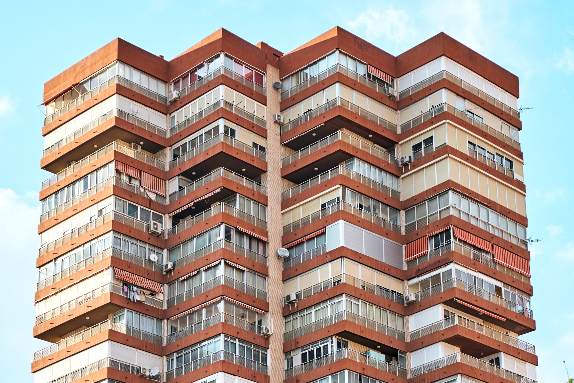 Imagem aproximada de um moderno edifício residencial de vários andares com fachada vermelha e bege contra um céu azul claro.
