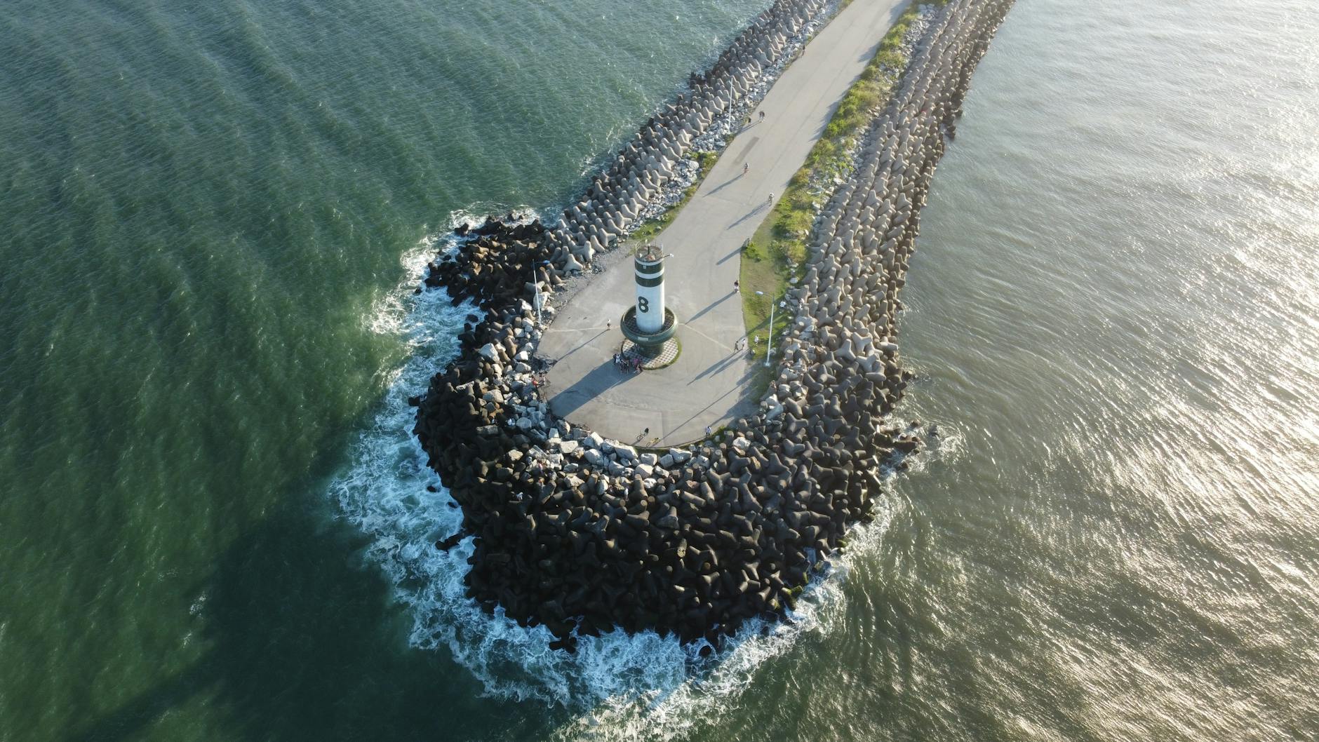 Vista aérea panorâmica do farol de Itajaí sobre um píer rochoso que se estende mar adentro, capturando as ondas e o oceano.